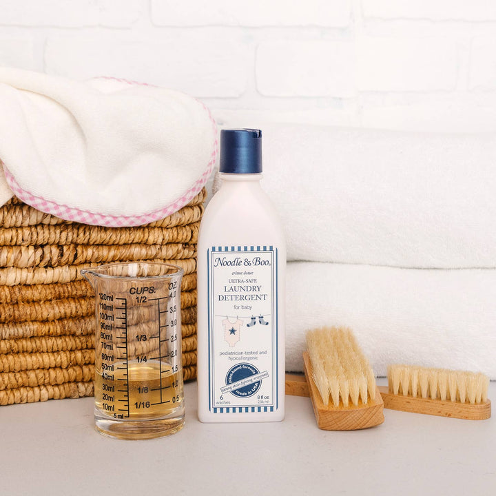 Bottle of baby laundry detergent with measuring cup, brush, and basket on a white background