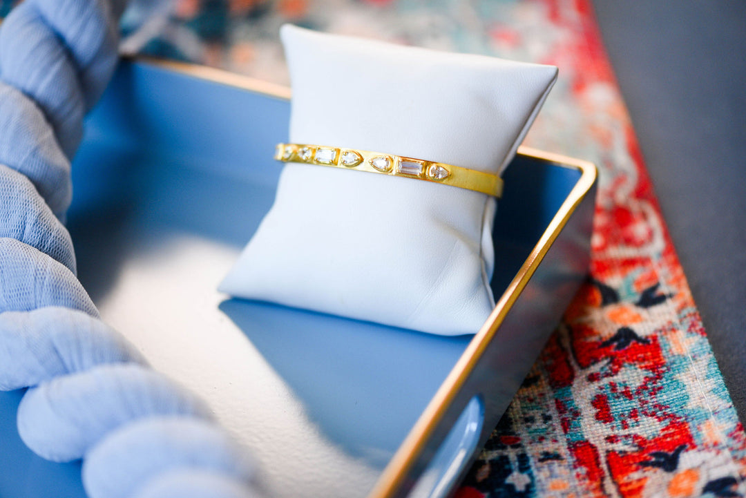 A gold bangle bracelet with clear stones, displayed on a white jewelry pillow inside a blue tray.