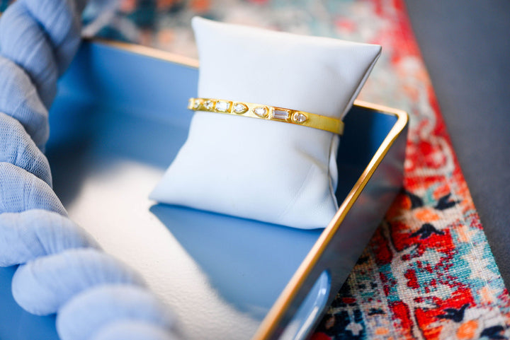 A gold bangle bracelet with clear stones, displayed on a white jewelry pillow inside a blue tray.