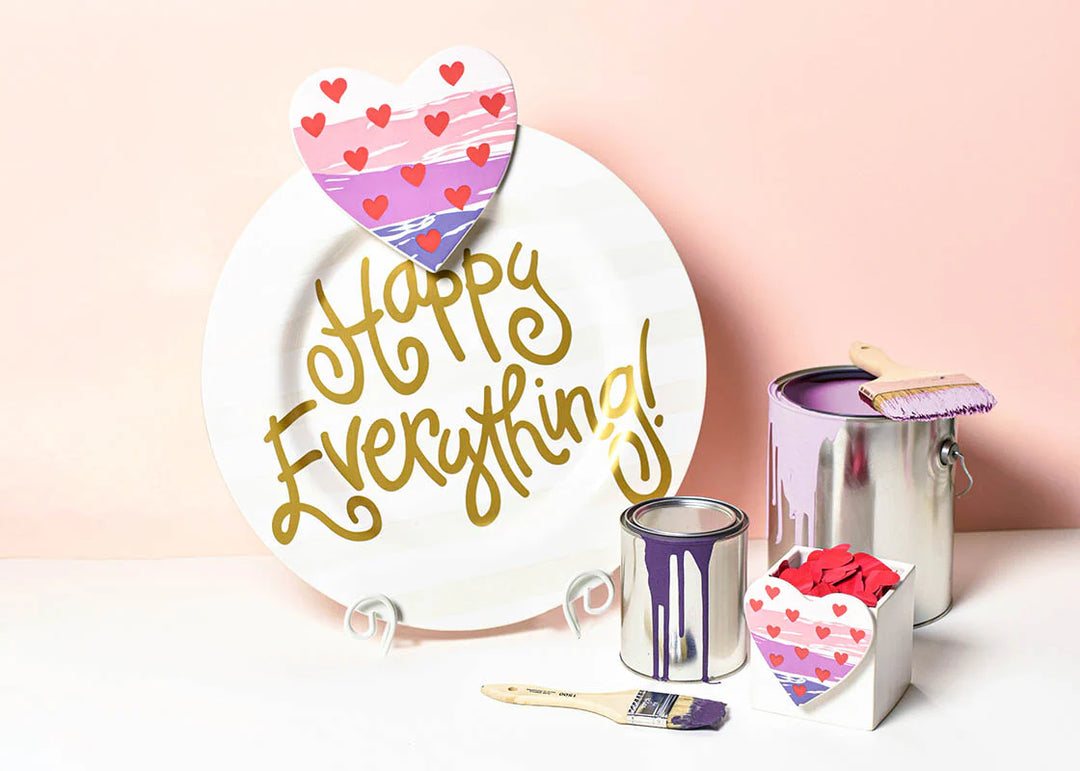 Decorative items including a heart-shaped sign, 'Happy Everything!' plate, and small paint cans on a light pink background.