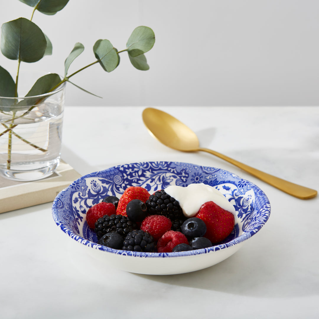 Bowl of berries with yogurt on a white surface with a gold spoon and plant in the background