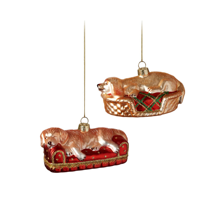 Two glass ornaments of dogs in a basket and on a red cushion with gold accents on a white background.