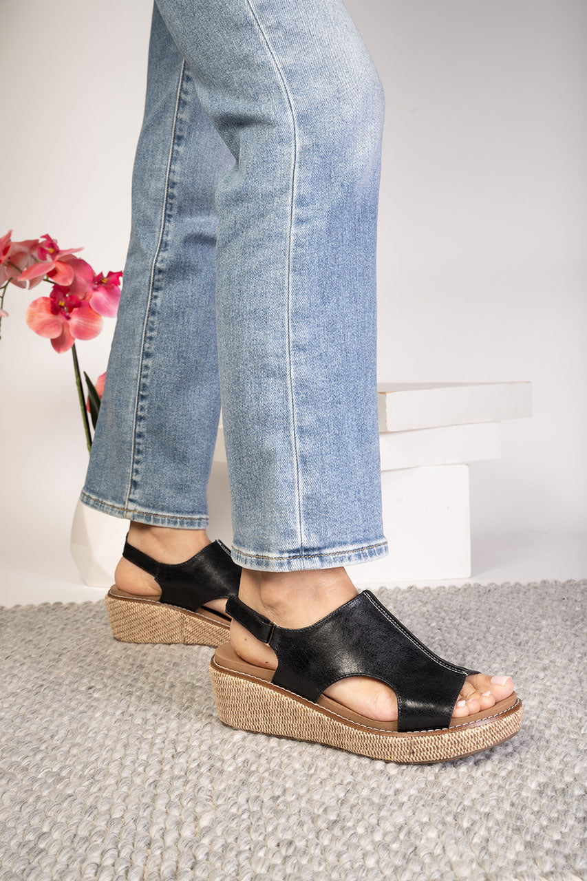 Person wearing black sandals with a textured sole, standing on a gray surface with a white wall and pink flowers in the background.