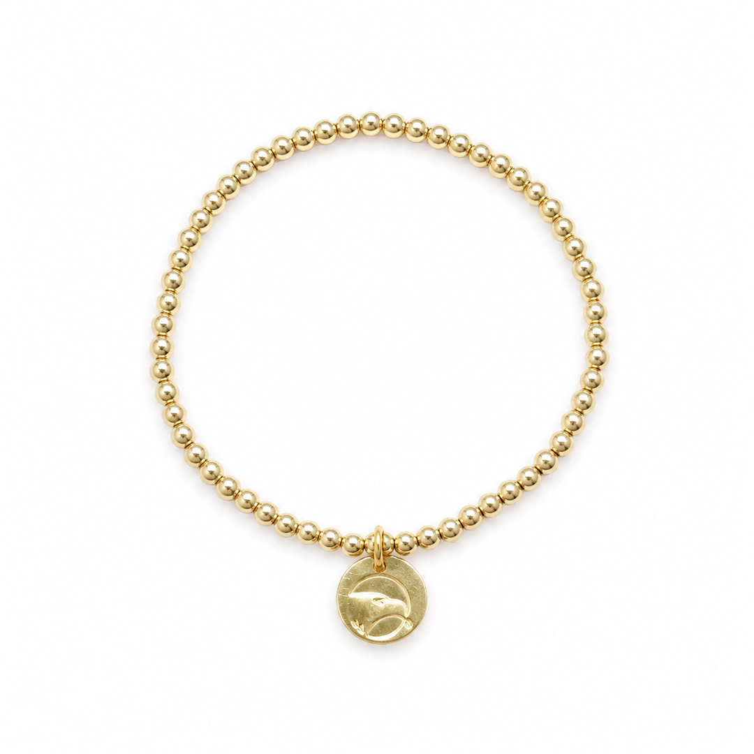 A gold beaded bracelet with a small round disc charm featuring a stylized eagle design, displayed on a clean white background.