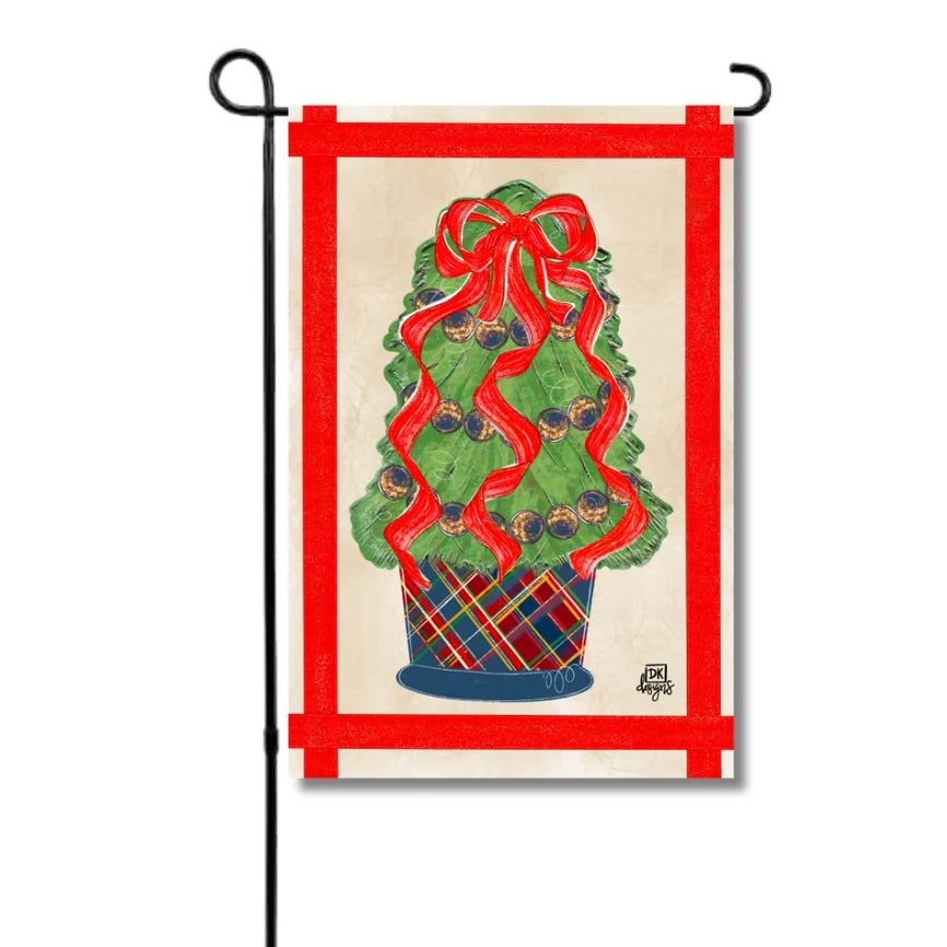 A vertical garden flag hanging from a black metal stand, featuring a decorated Christmas tree in a plaid pot with a large red ribbon bow, set against a cream background with a red border.