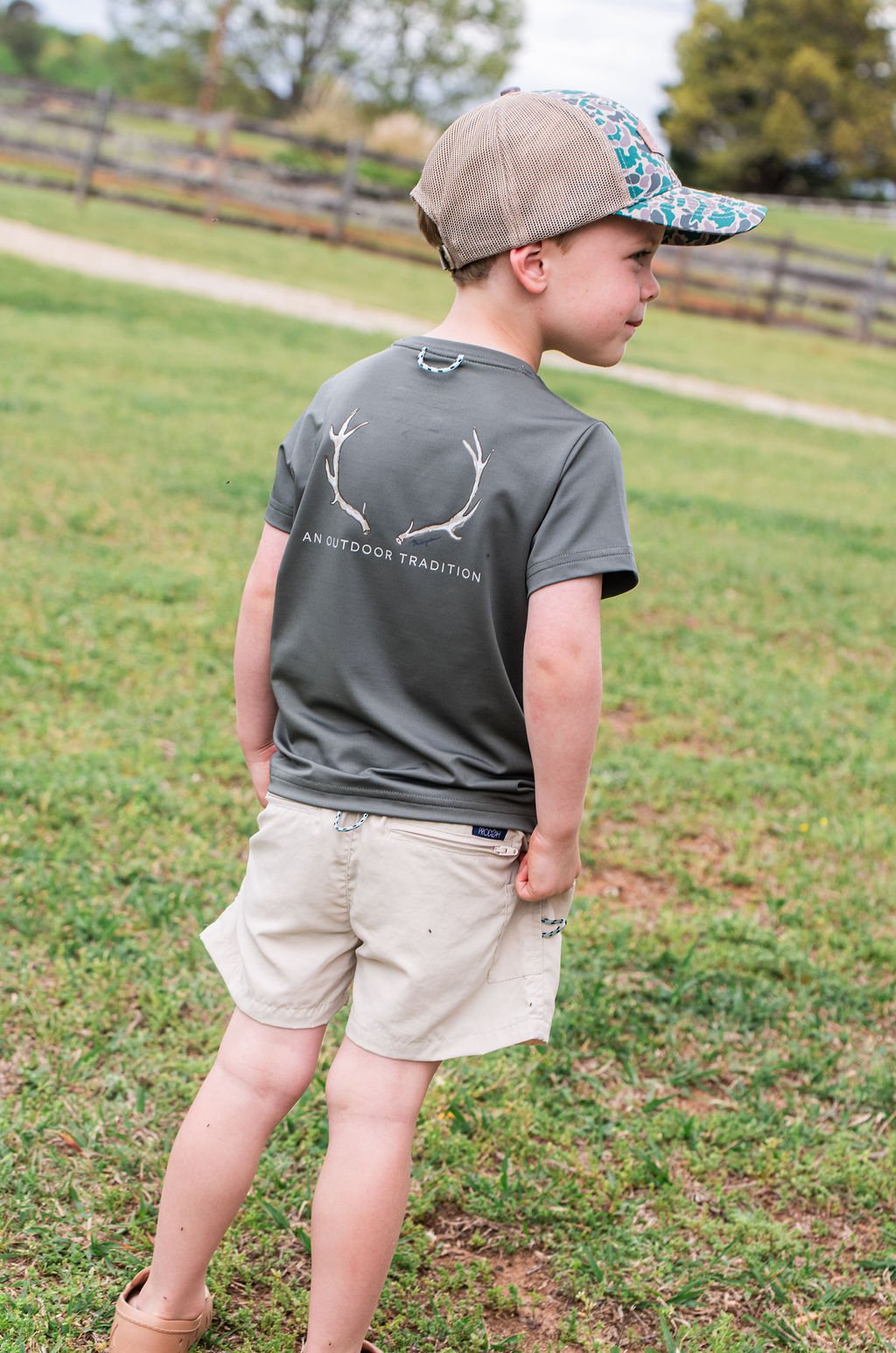 Child wearing a gray t-shirt with a logo and beige shorts standing in a grassy field.
