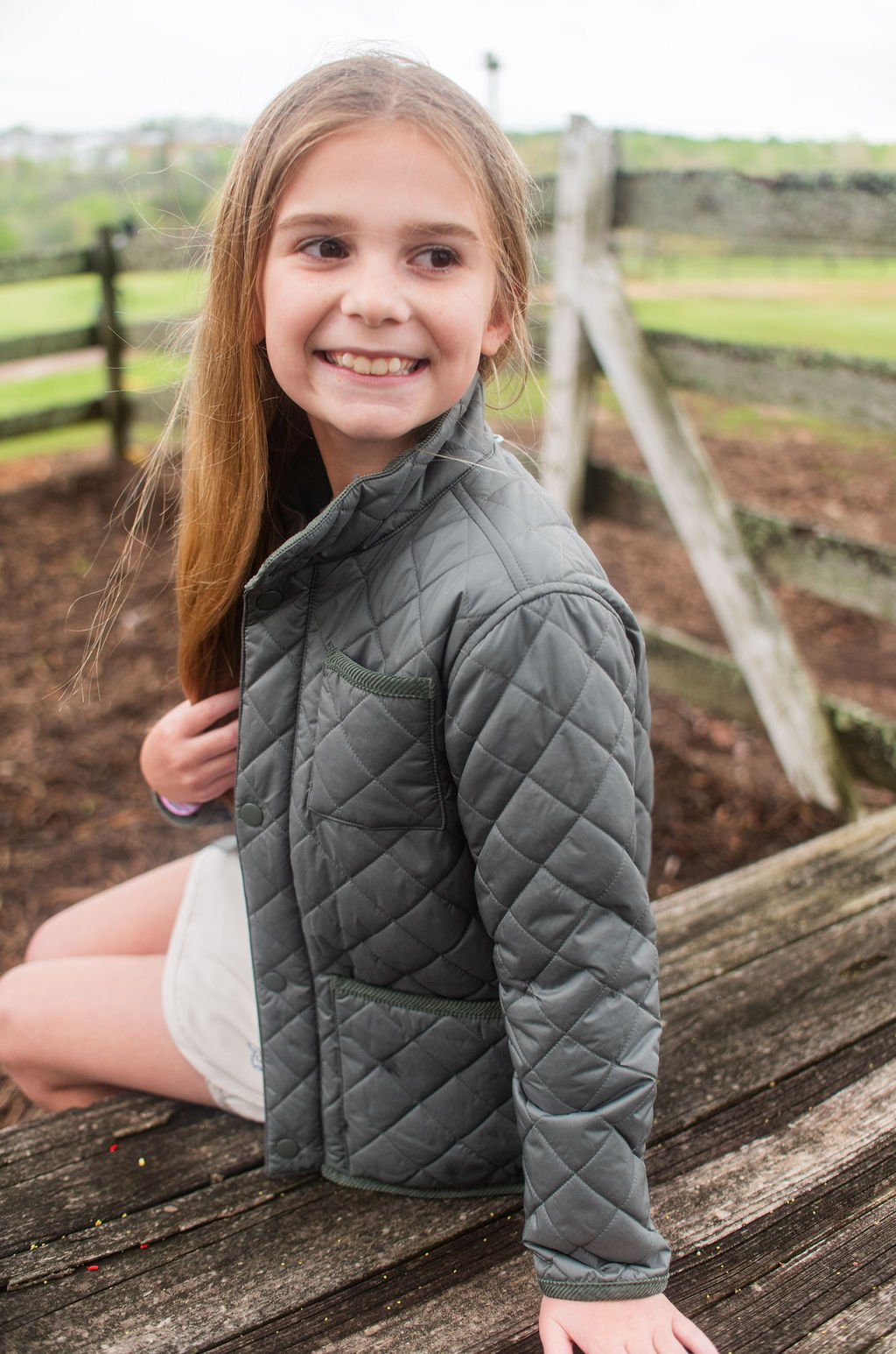 Young girl wearing a dark green quilted jacket sitting on a wooden bench outdoors.