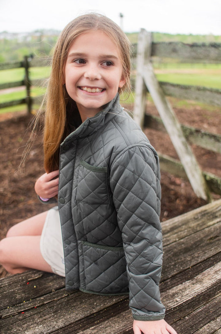 Young girl wearing a dark green quilted jacket sitting on a wooden bench outdoors.