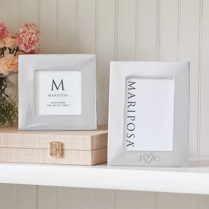 Two silver picture frames displayed on a white shelf, one standing upright on a beige textured box and the other lying flat. The frames have white mat borders with custom monogram designs, and pink flowers are visible in the background against a white paneled wall.