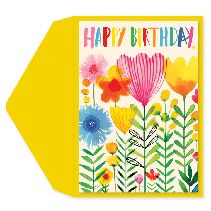 A square birthday card with a bright yellow envelope behind it. The card has a cream background with colorful floral illustrations and the words “HAPPY BIRTHDAY” in multicolored letters at the top.