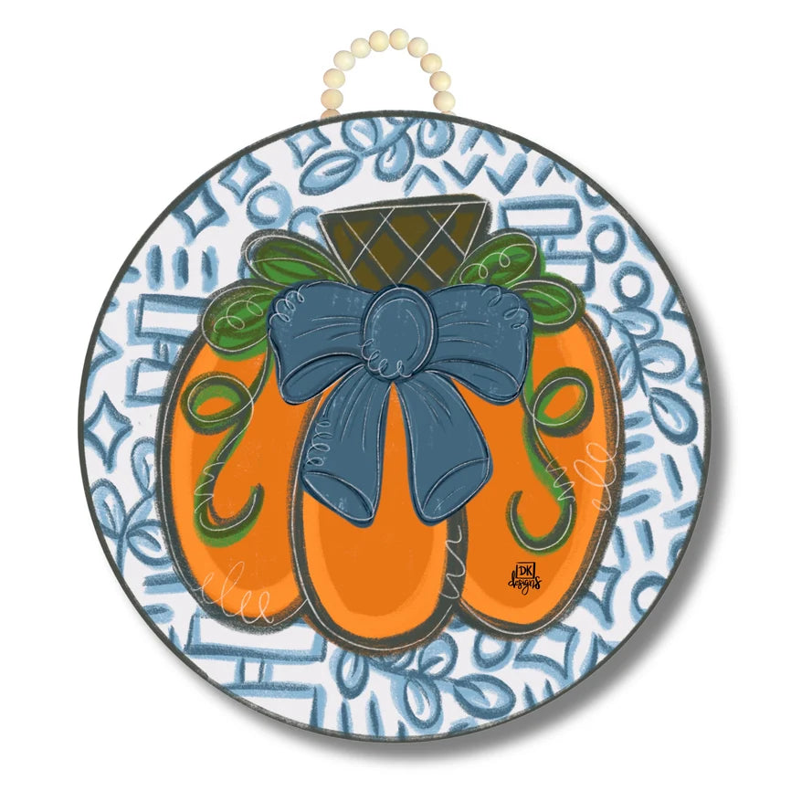 A round decorative wall hanging featuring a stylized orange pumpkin with a large blue bow, green leaves, and a patterned blue and white background, suspended by a cream-colored beaded loop at the top.