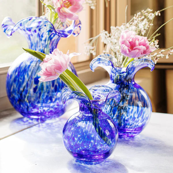 Three blue glass vases with floral arrangements on a reflective surface.
