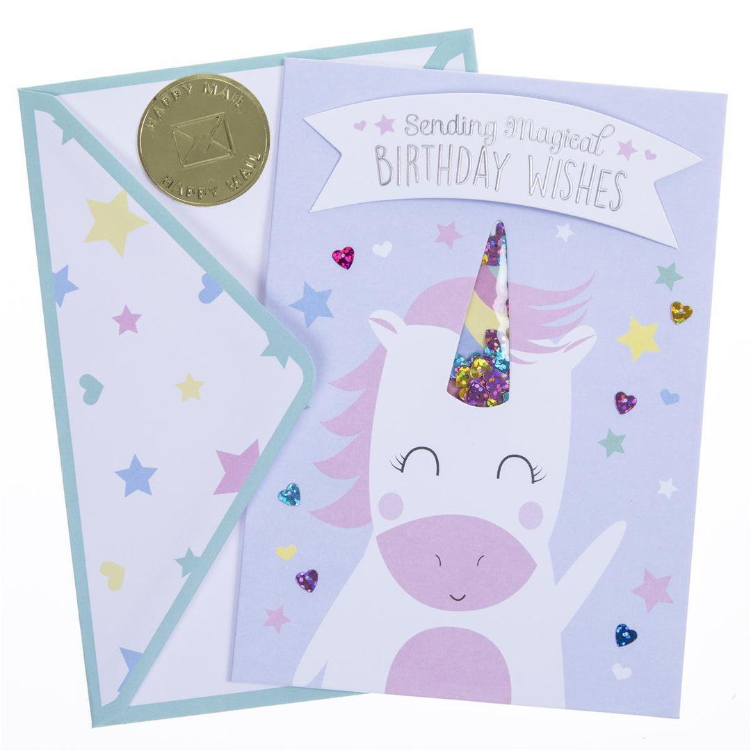 A pastel unicorn birthday card with a matching envelope. The card features a white unicorn with a pink snout and mane, a colorful glitter-filled horn, and a banner reading 'Sending Magical Birthday Wishes.