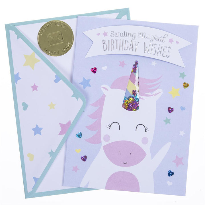 A pastel unicorn birthday card with a matching envelope. The card features a white unicorn with a pink snout and mane, a colorful glitter-filled horn, and a banner reading 'Sending Magical Birthday Wishes.