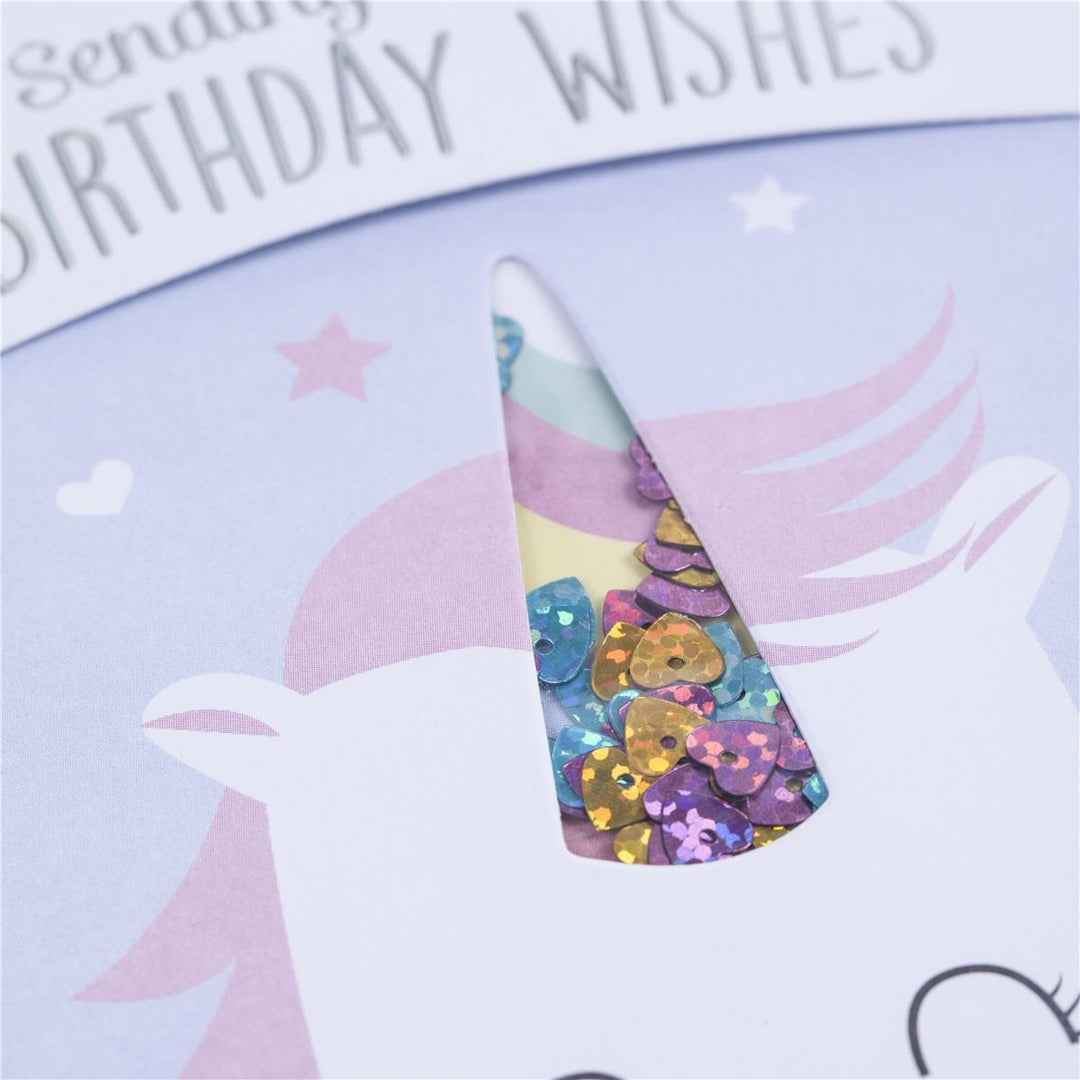 A close-up of a unicorn-themed birthday card with a die-cut window showing a colorful party hat filled with glittering sequins. The card has a pastel purple background with a white unicorn illustration and the text 'Sending Birthday Wishes' at the top.