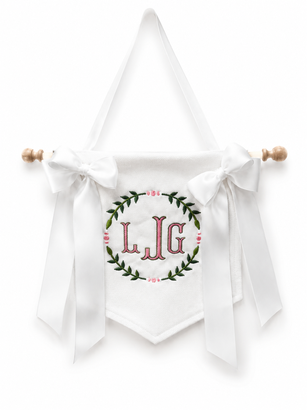 A white fabric baby bib or pouch with a long hanging ribbon, bow, and wooden dowel, featuring an embroidered green wreath and pink initials 'L.J.