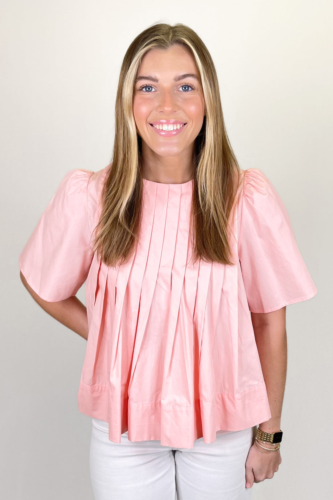 Woman modeling a peach pleated blouse with short flutter sleeves paired with white pants.