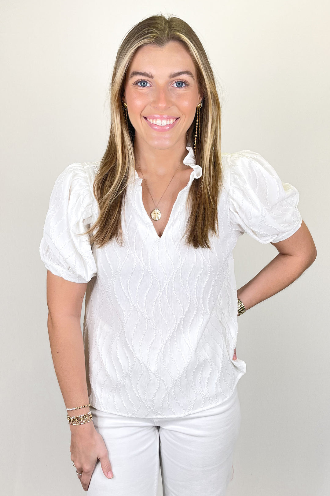 Woman modeling a white textured blouse with puff sleeves and split ruffle neckline paired with white pants.