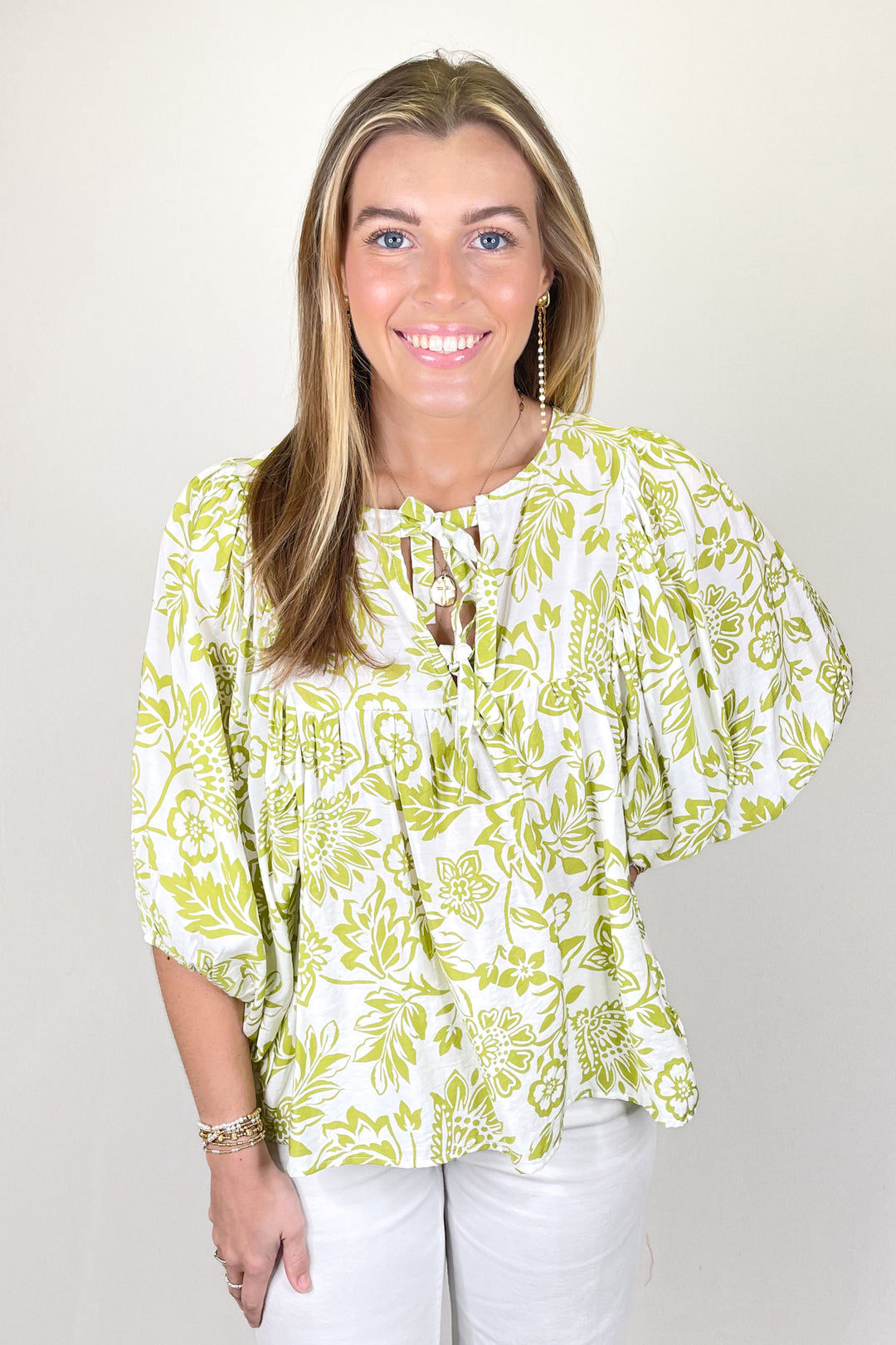 Woman modeling a green and white floral print blouse with tie neckline and three-quarter sleeves paired with white pants.