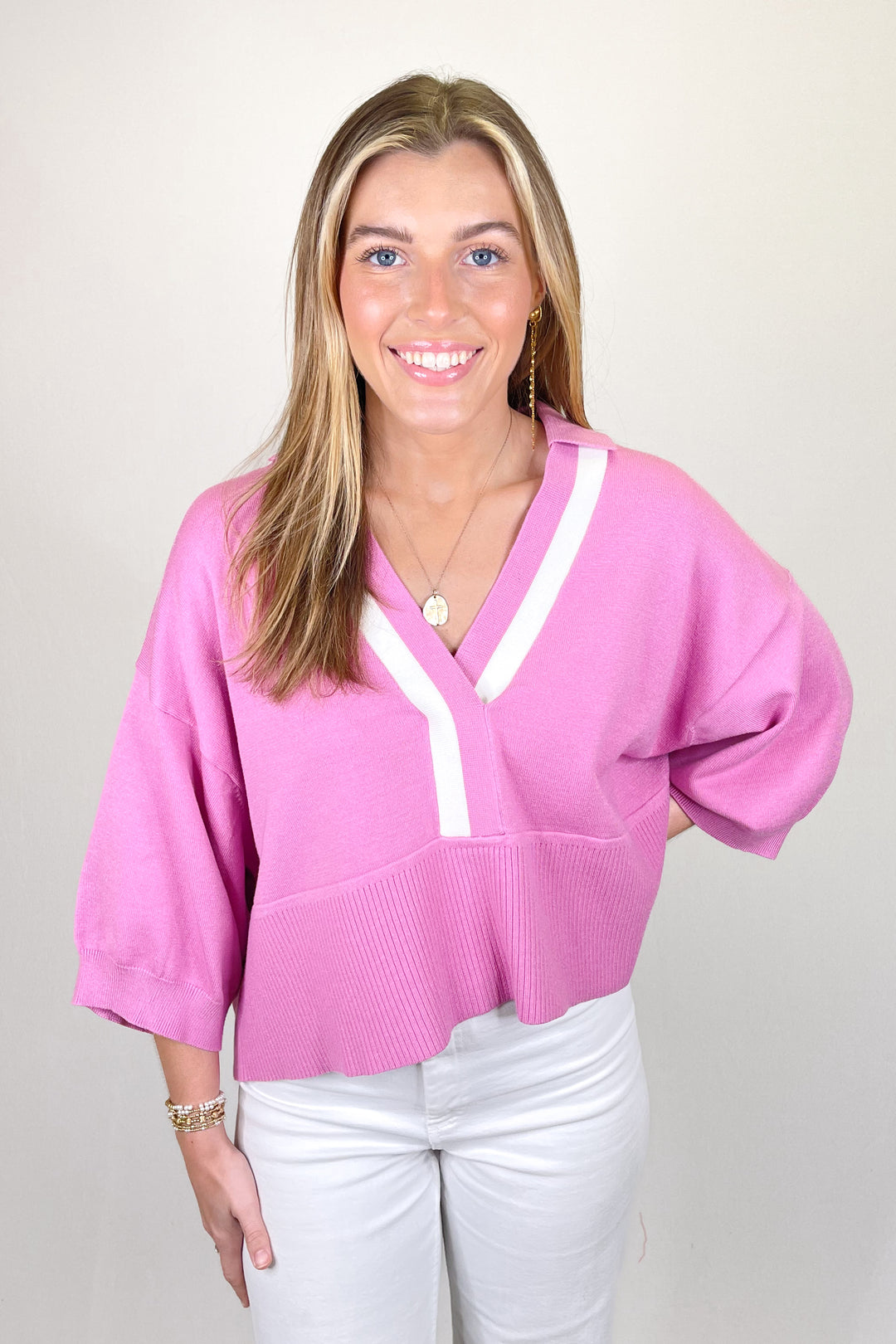 Woman modeling a pink knit short sleeve sweater with white contrast V-neck detail paired with white pants.