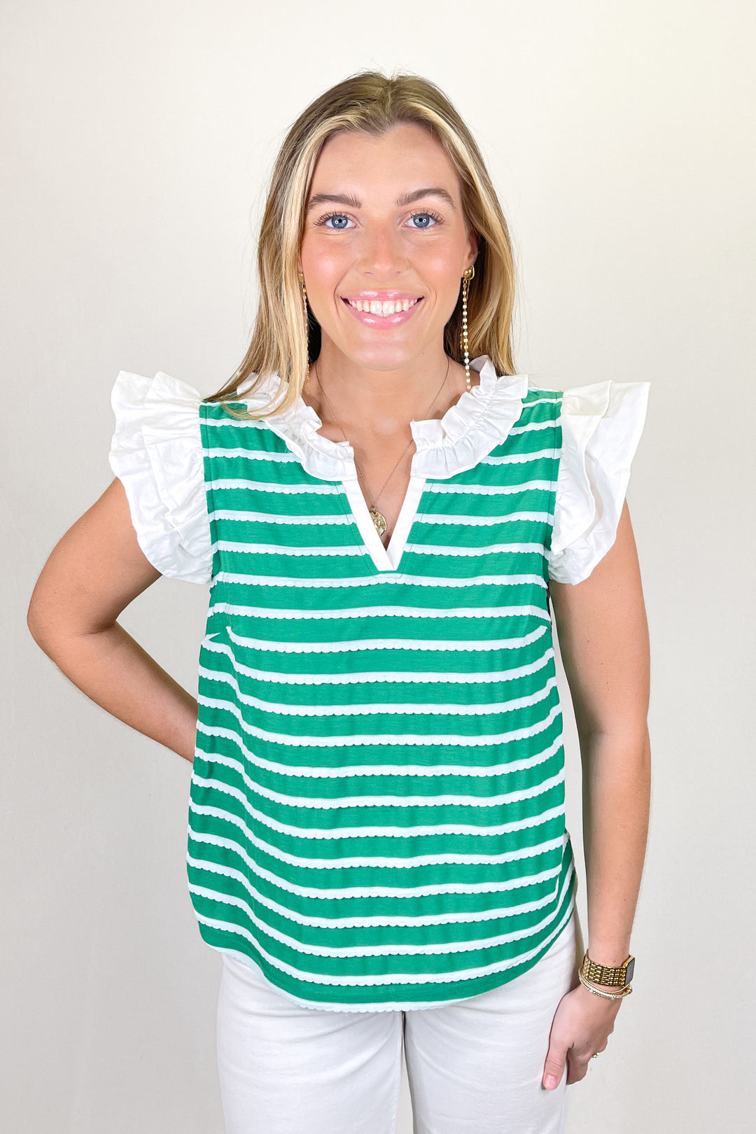 Woman modeling a green and white striped sleeveless blouse with ruffle shoulders and split neckline paired with white pants.