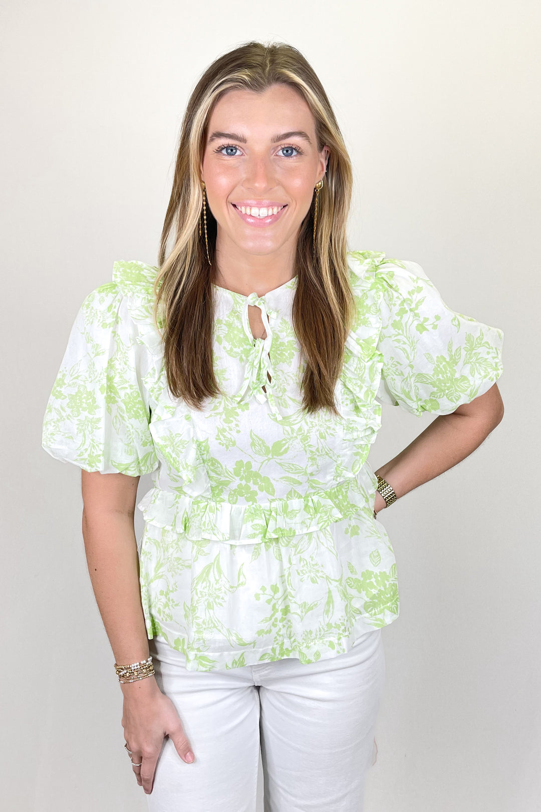 Woman modeling a white and light green floral blouse with ruffle details and short puff sleeves paired with white pants.
