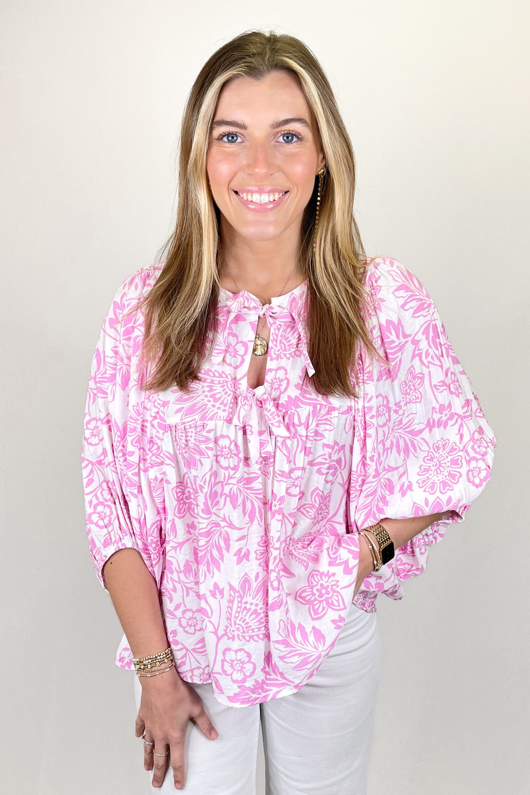 Woman modeling a pink and white floral print blouse with tie neckline and three-quarter sleeves paired with white pants.