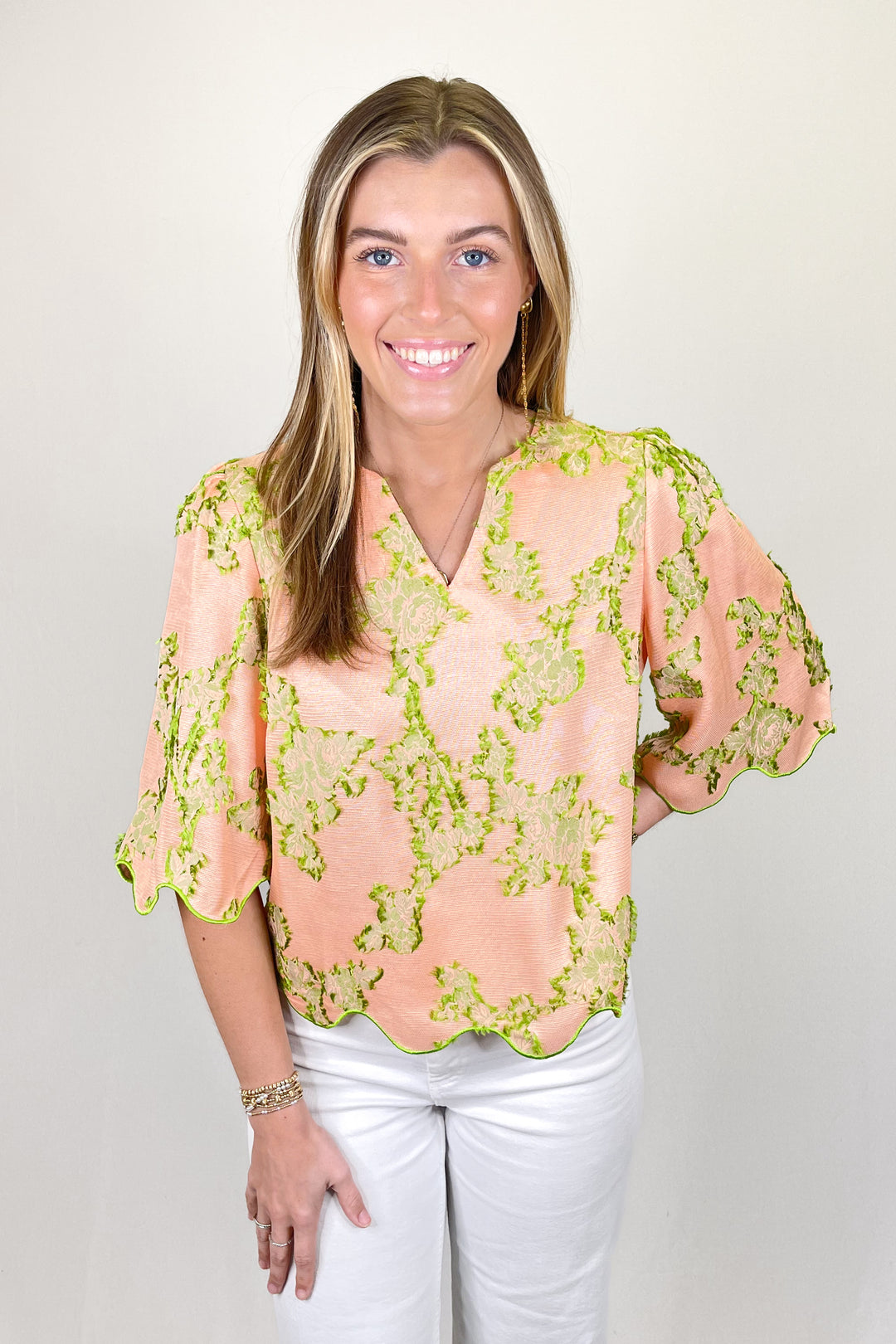 Woman modeling a peach blouse with green floral embroidery, scalloped hem, and flutter sleeves paired with white pants.