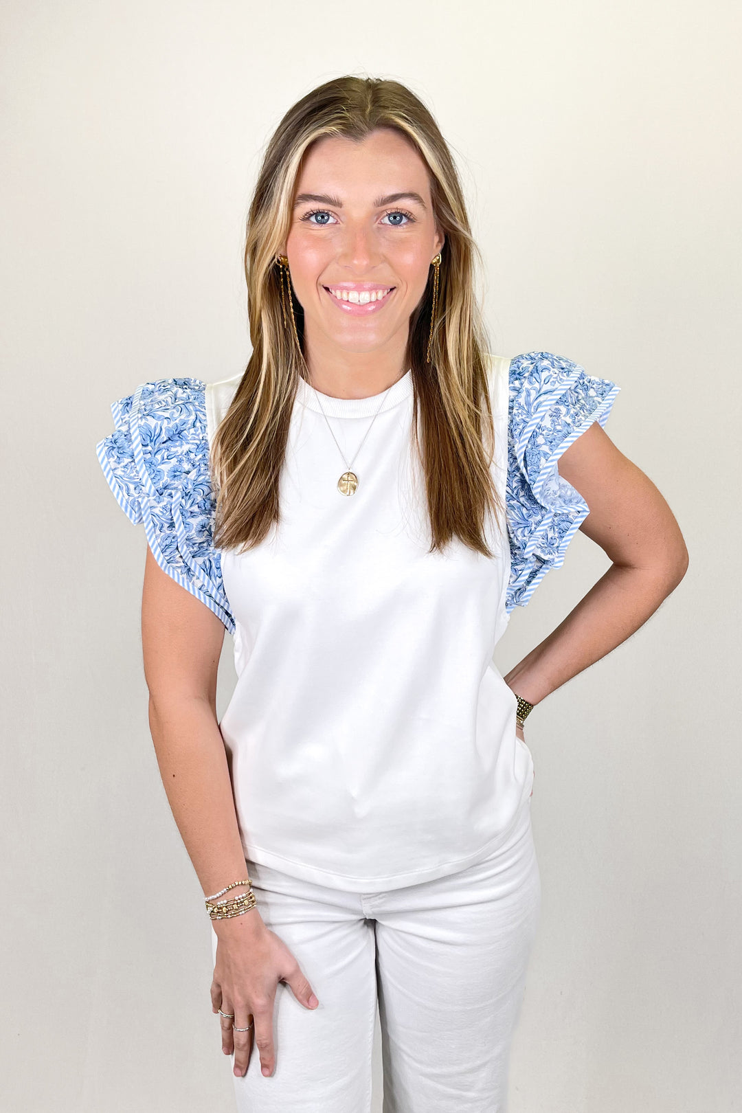 Woman modeling a white sleeveless top with blue floral ruffle shoulder detail paired with white pants.