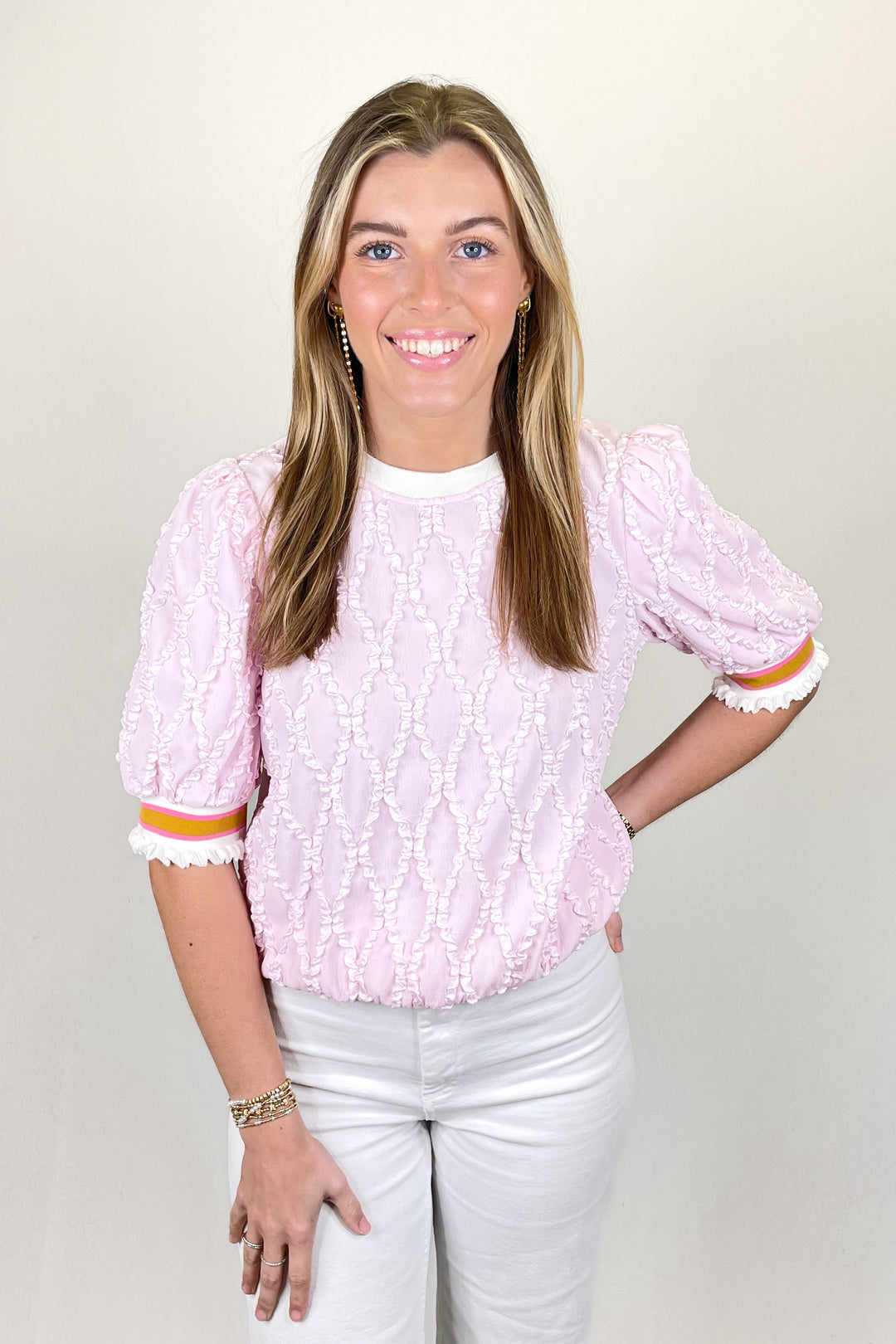 Woman modeling a light pink textured blouse with puff sleeves and striped cuff detail paired with white pants.