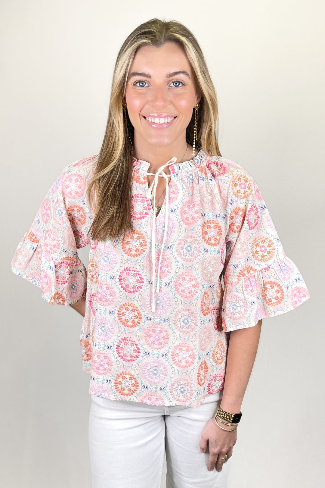 Woman modeling a pastel floral print blouse with tie neckline and short ruffle sleeves paired with white pants.