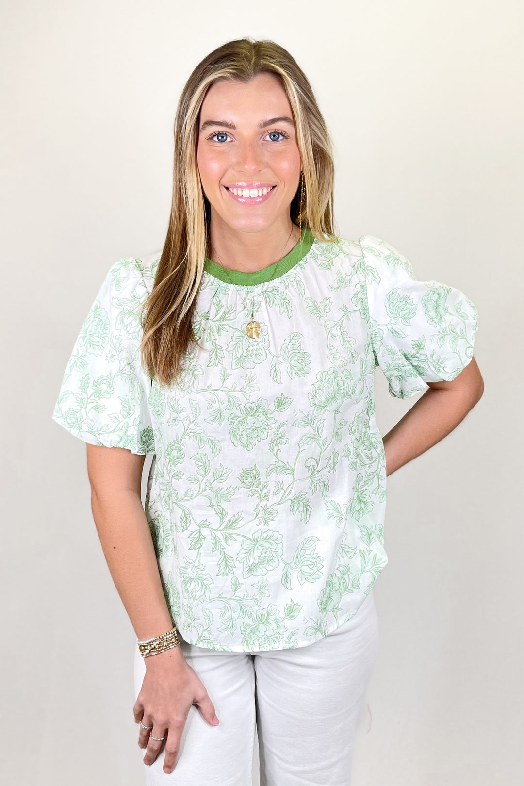 Woman modeling a white and light green floral blouse with puff sleeves and green trim neckline paired with white pants.