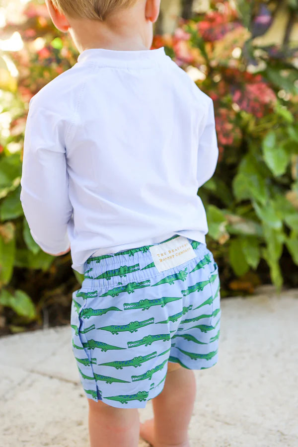 Child wearing a light blue long-sleeve shirt and green alligator shorts with a floral background.