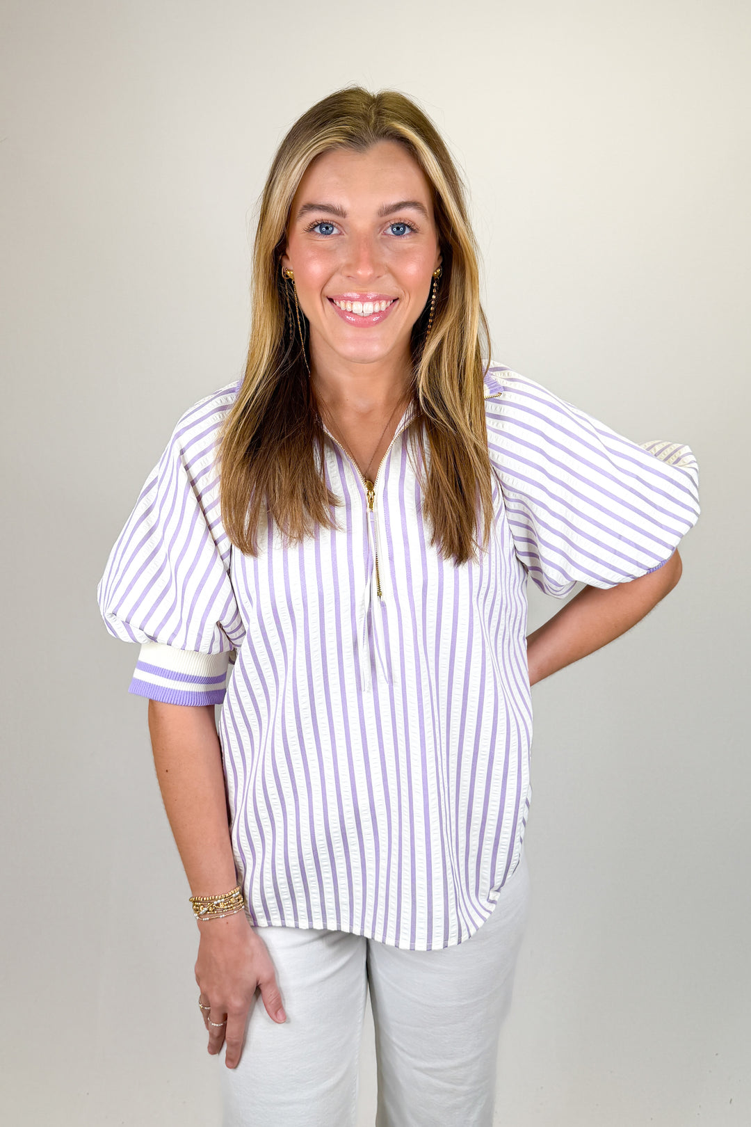 Woman modeling a lavender and white striped blouse with puff sleeves and half-zip neckline paired with white pants.