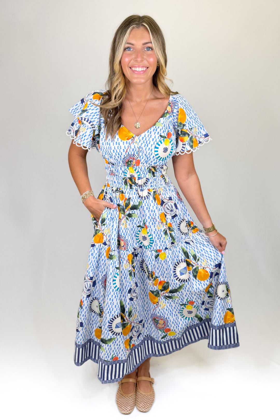 Blue and white printed midi dress with flutter sleeves, V-neckline, smocked waist, and tiered skirt, featuring a citrus and coastal-inspired print with contrast border detail
