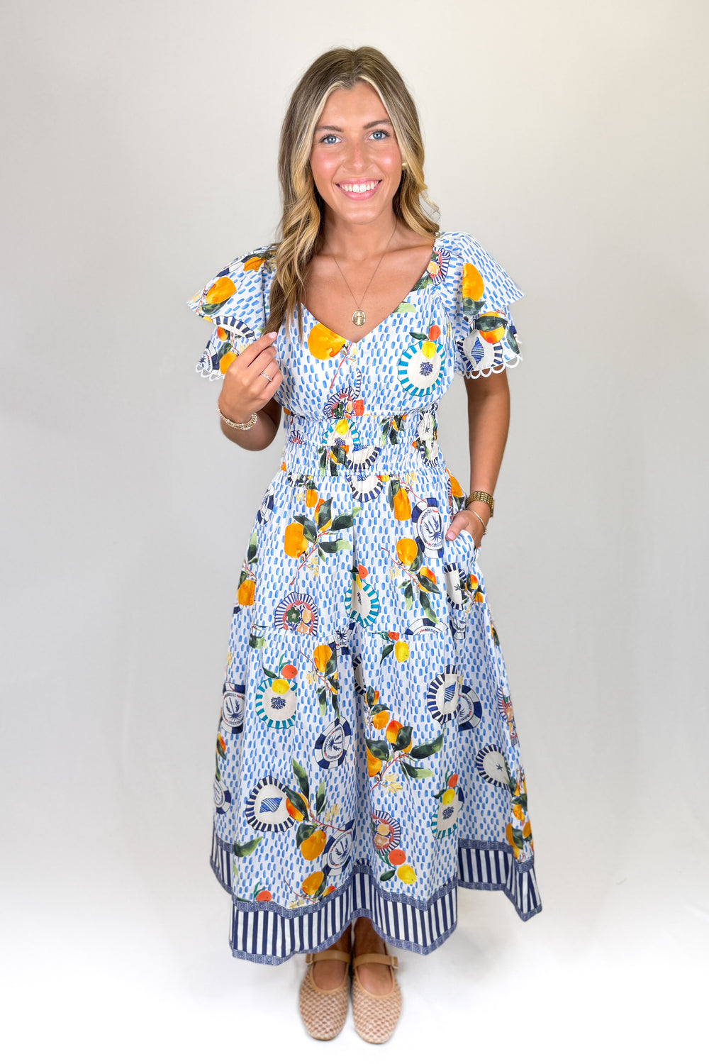 Blue and white printed midi dress with flutter sleeves, V-neckline, smocked waist, and tiered skirt, featuring a citrus and coastal-inspired print with contrast border detail