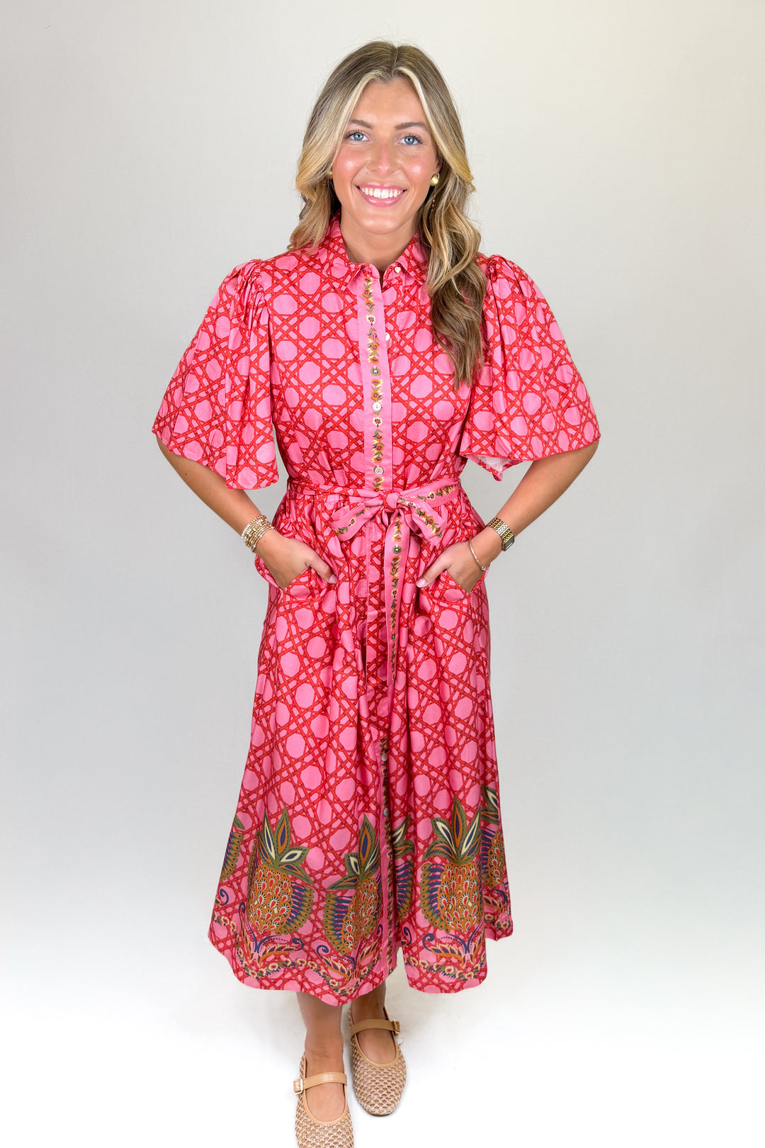 Pink patterned midi dress with short puff sleeves, collared neckline, button front, and tie waist, featuring a contrast border hem, pockets, and a flowy fit
