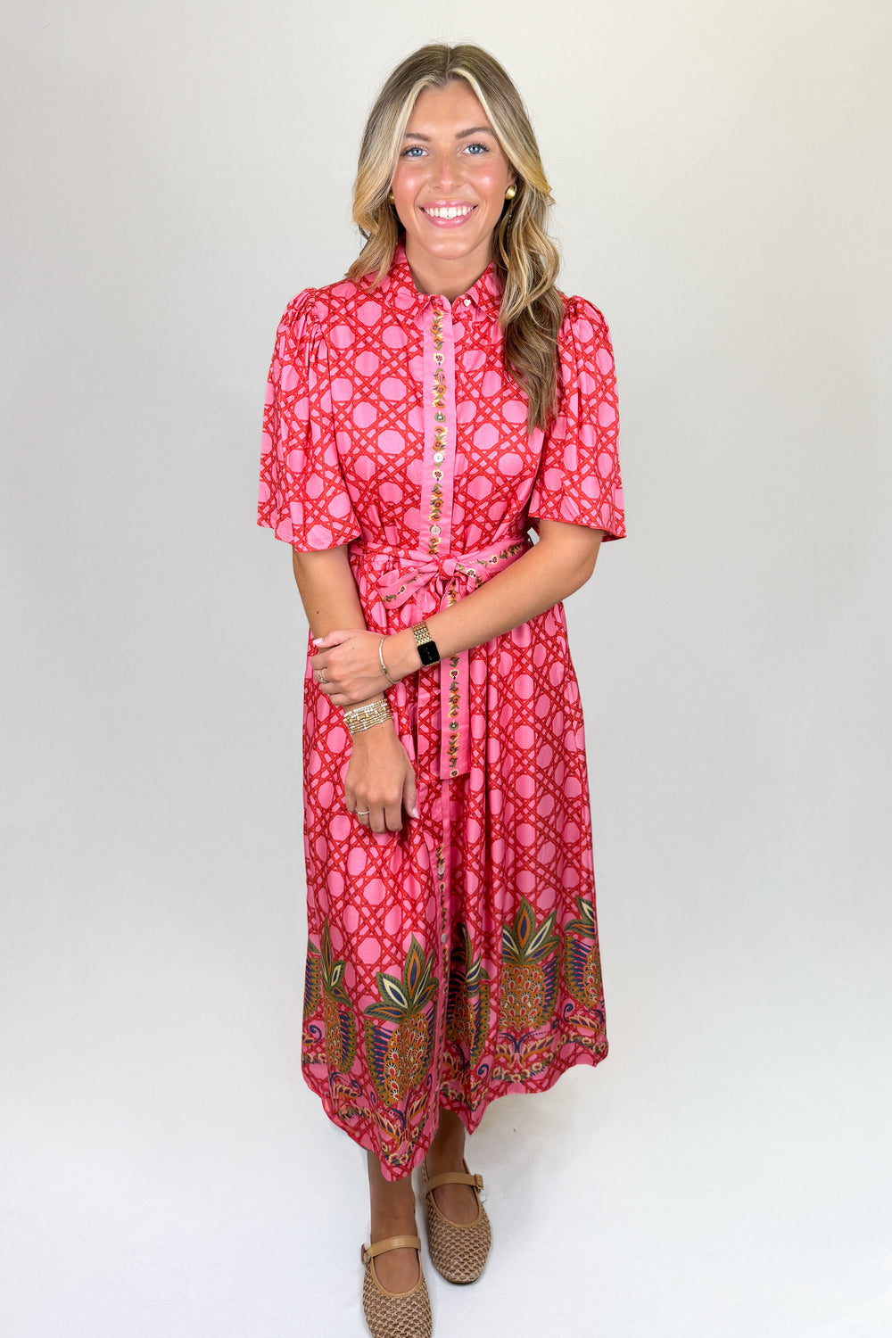 Pink patterned midi dress with short puff sleeves, collared neckline, button front, and tie waist, featuring a contrast border hem, pockets, and a flowy fit