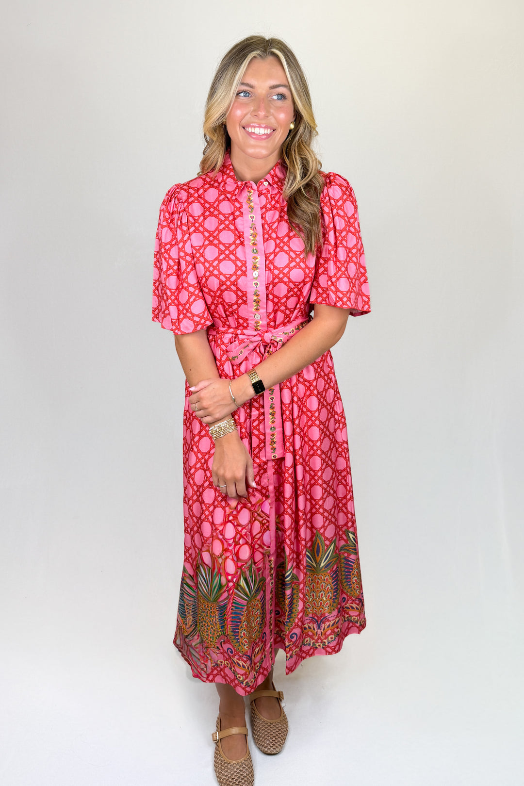 Pink patterned midi dress with short puff sleeves, collared neckline, button front, and tie waist, featuring a contrast border hem, pockets, and a flowy fit