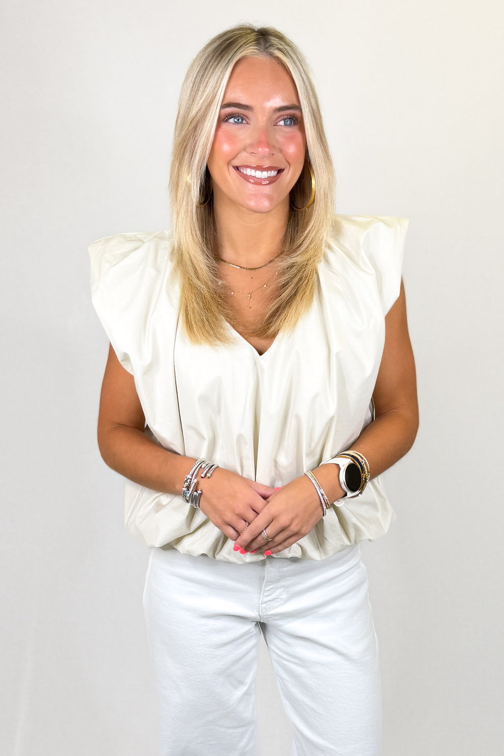Cream sleeveless bubble hem blouse with structured ruffle shoulders paired with white denim and layered gold jewelry for a chic neutral summer outfit.