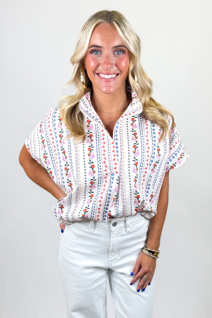 Woman wearing a patterned blouse and light pants against a plain background