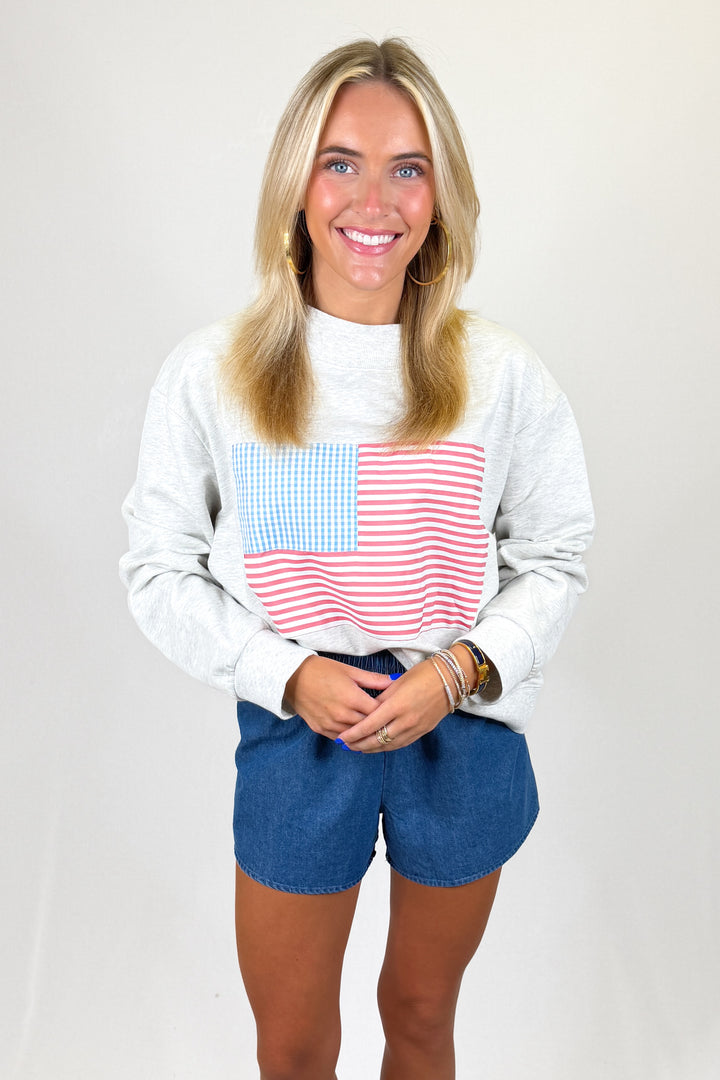 Woman wearing a white sweatshirt with an American flag design and blue shorts on a plain background