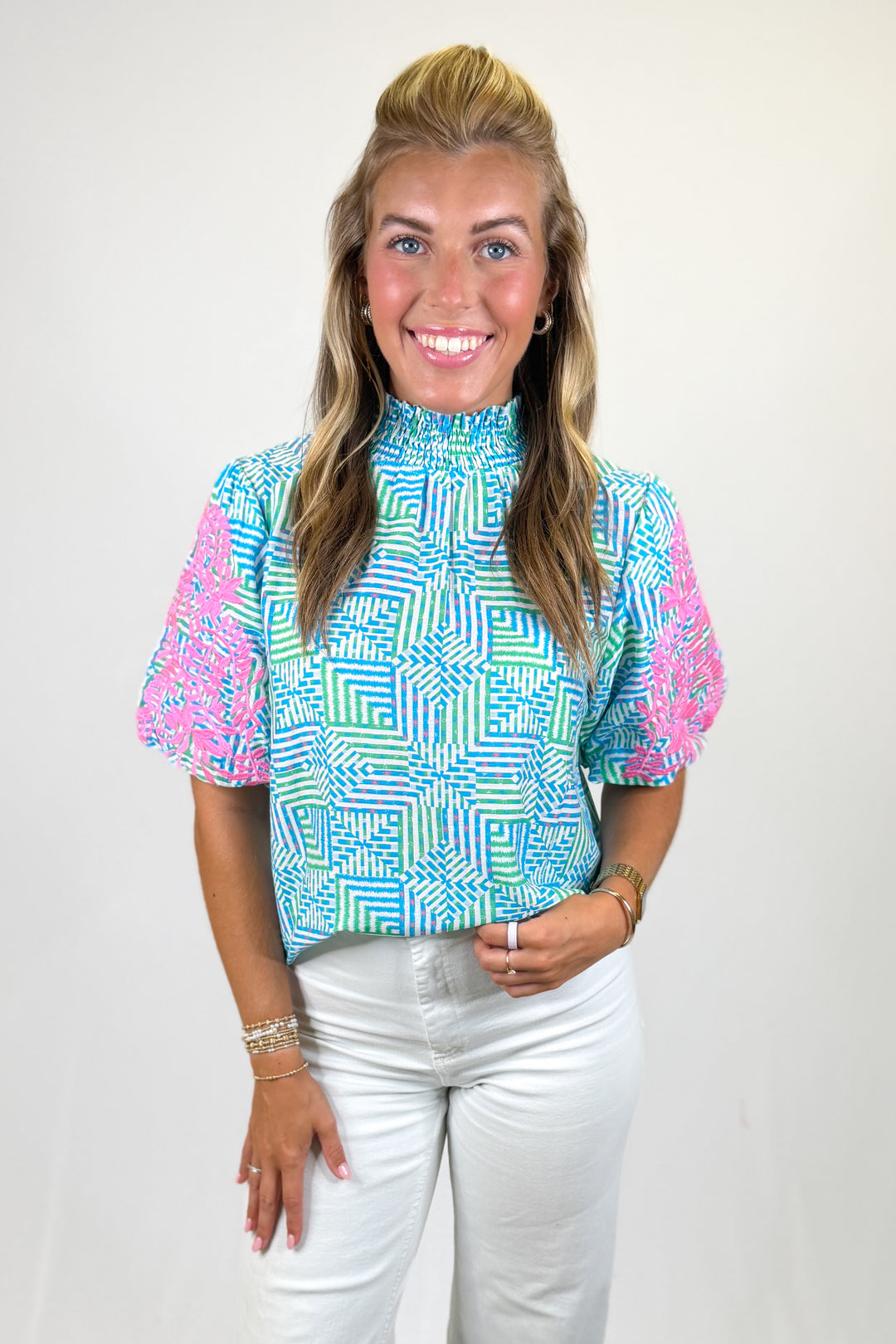 Woman wearing a colorful geometric patterned top with white pants on a plain background