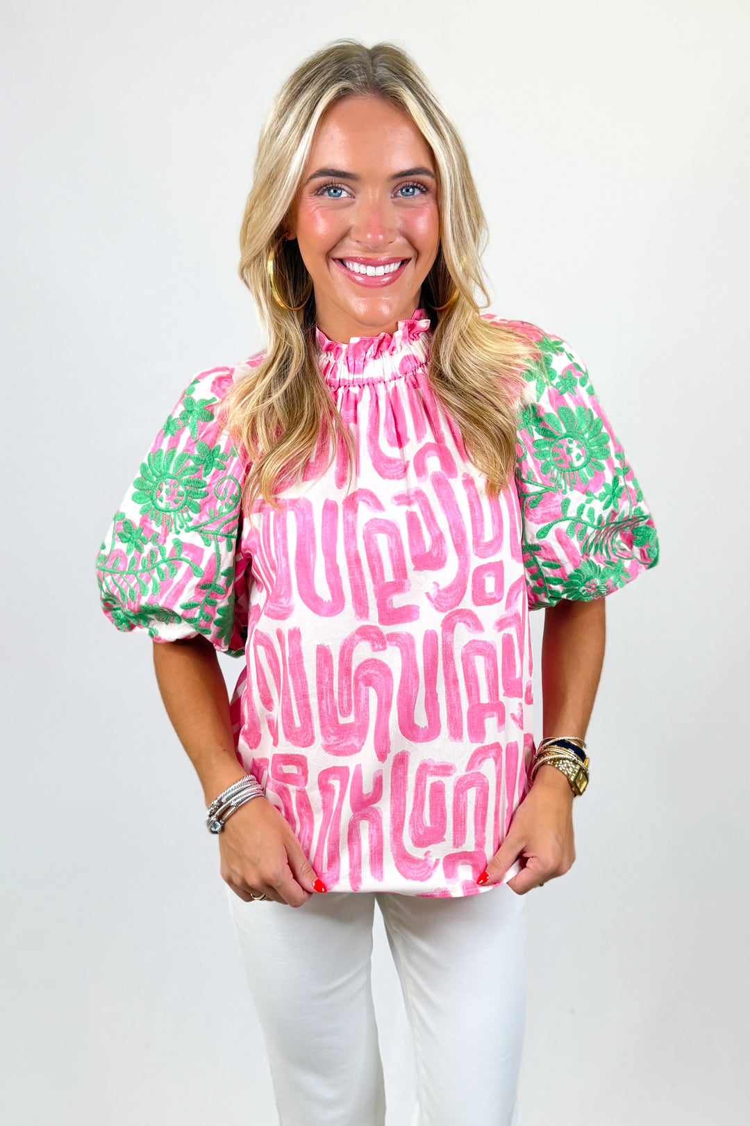 Woman wearing a pink and green patterned blouse with white pants on a plain background
