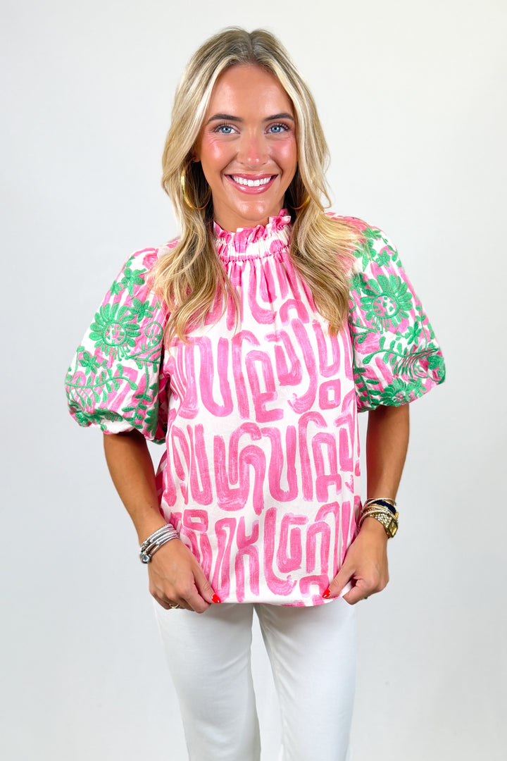 Woman wearing a pink and green patterned blouse with white pants on a plain background