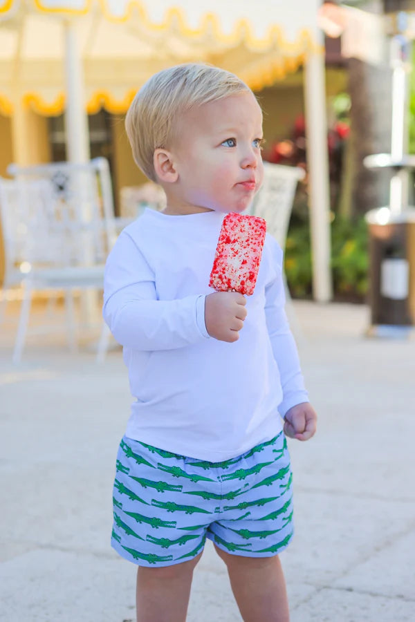 Child wearing a white long-sleeve shirt and green shorts with alligator pattern, holding a red popsicle.