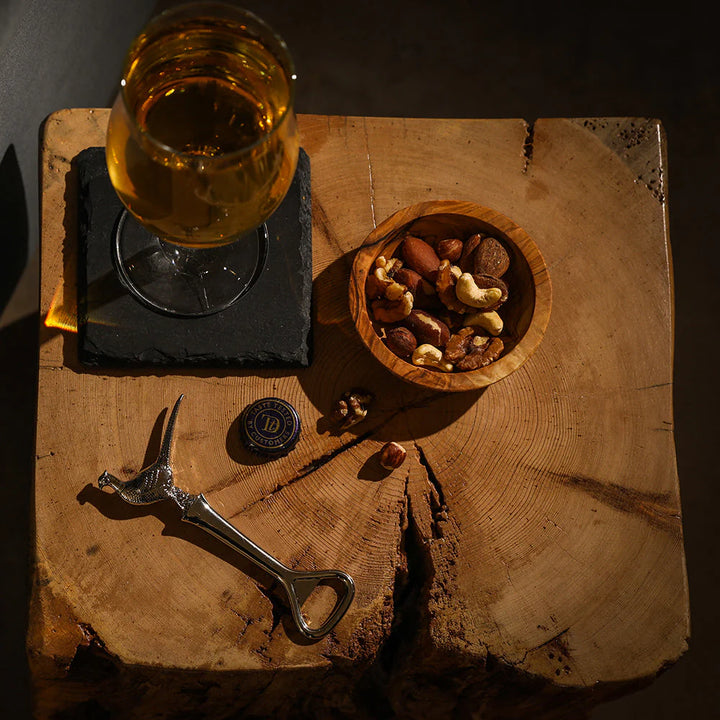 Glass of beer, bowl of nuts, and bottle opener on a wooden surface