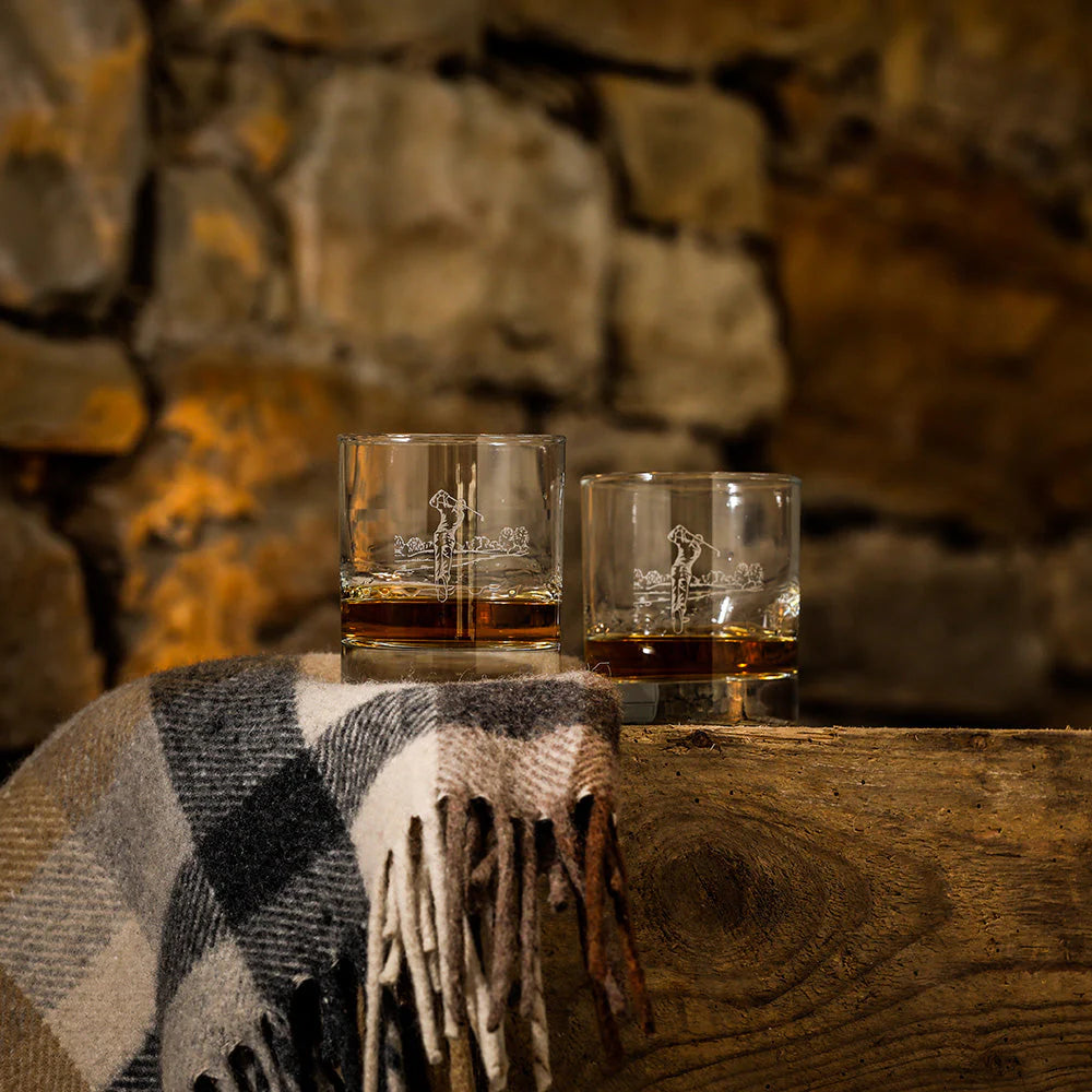 Two glasses of whiskey on a wooden surface with a stone wall background
