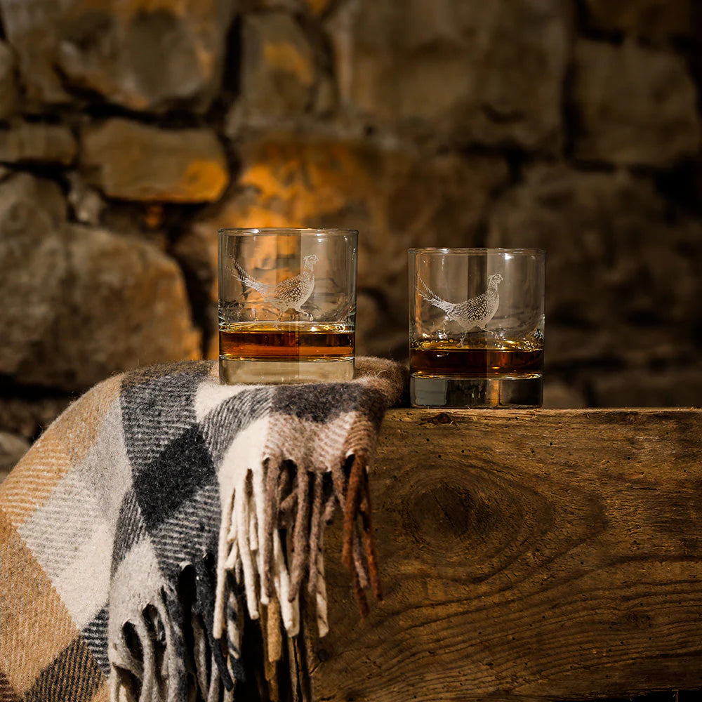 Two glasses of whiskey on a wooden surface with a stone wall background