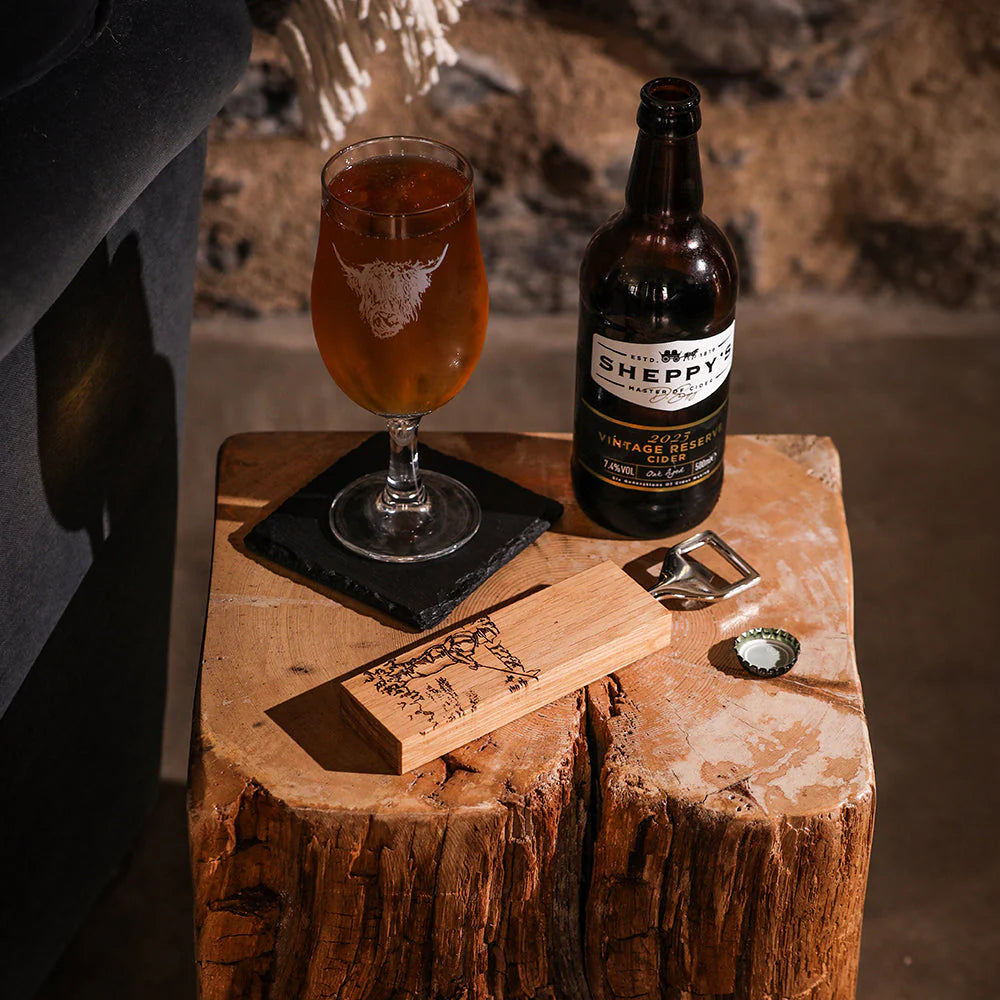 Sheppy beer bottle with a glass of amber ale on a wooden stool by a fireplace.